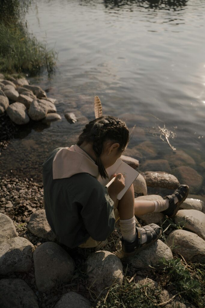 A girl with braided hair sits by a lake, drawing in a notebook. Serene outdoor setting.